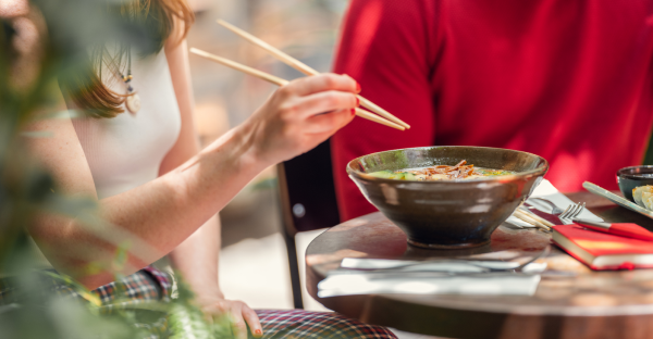 Two people sitting at a table with one eating a bowl of ramen with chopsticks